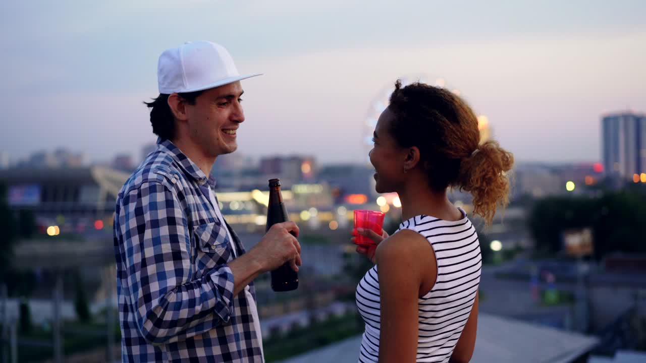 Giovane coppia multietnica, ragazza afroamericana e ragazzo caucasico, stanno bevendo e parlando in piedi insieme sul tetto con una bellissima città sullo sfondo.