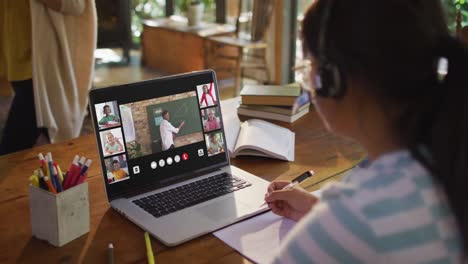 Schoolgirl-using-laptop-for-online-lesson-at-home,-with-diverse-teacher-and-class-on-screen