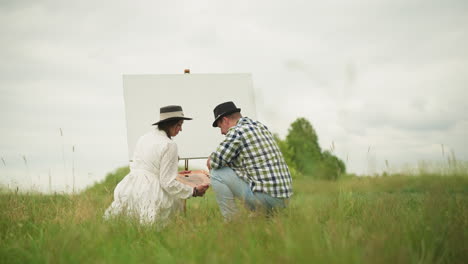 a man and a woman, both bent over in a grassy field, focused on their artistic tasks. the woman in a white dress holds a palette, while the man in a checked shirt and hat appears to be carefully