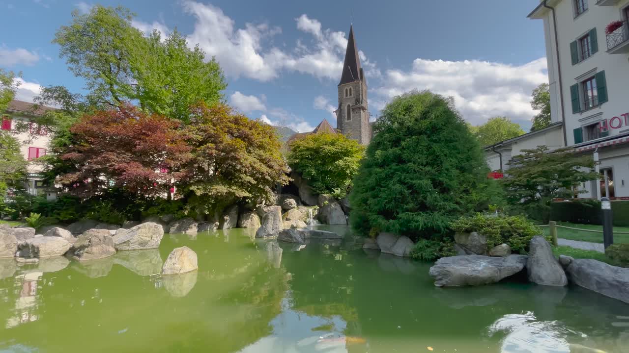 スイスのインターラーケンにある、緑豊かな鯉の池、岩、青々とした木々、そして教会の尖塔が配された静謐な日本庭園の静止画。空は一部曇り。