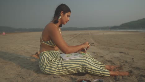 Indian-Woman-Art-Painter-Sitting-in-Sand-Puts-Paint-on-Her-Palette,-Side-Angle