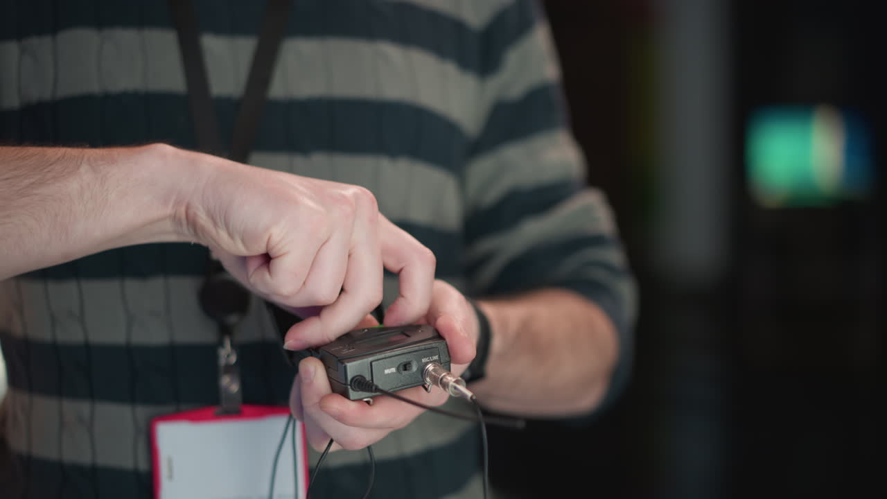 close-up of man adjusting wireless audio transmitter in hands, with cable attached, wearing striped sweater and lanyard with ID badge, preparing equipment for video production in studio setting