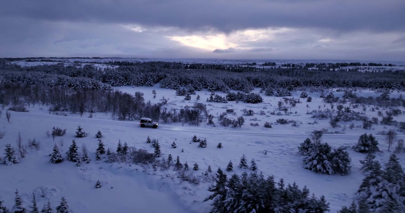 Drone sidebilde av en hvit SUV som kjører over en snødekt islandsk vei mellom rimdekte trær og lavafelt under en glødende vinterhimmel.