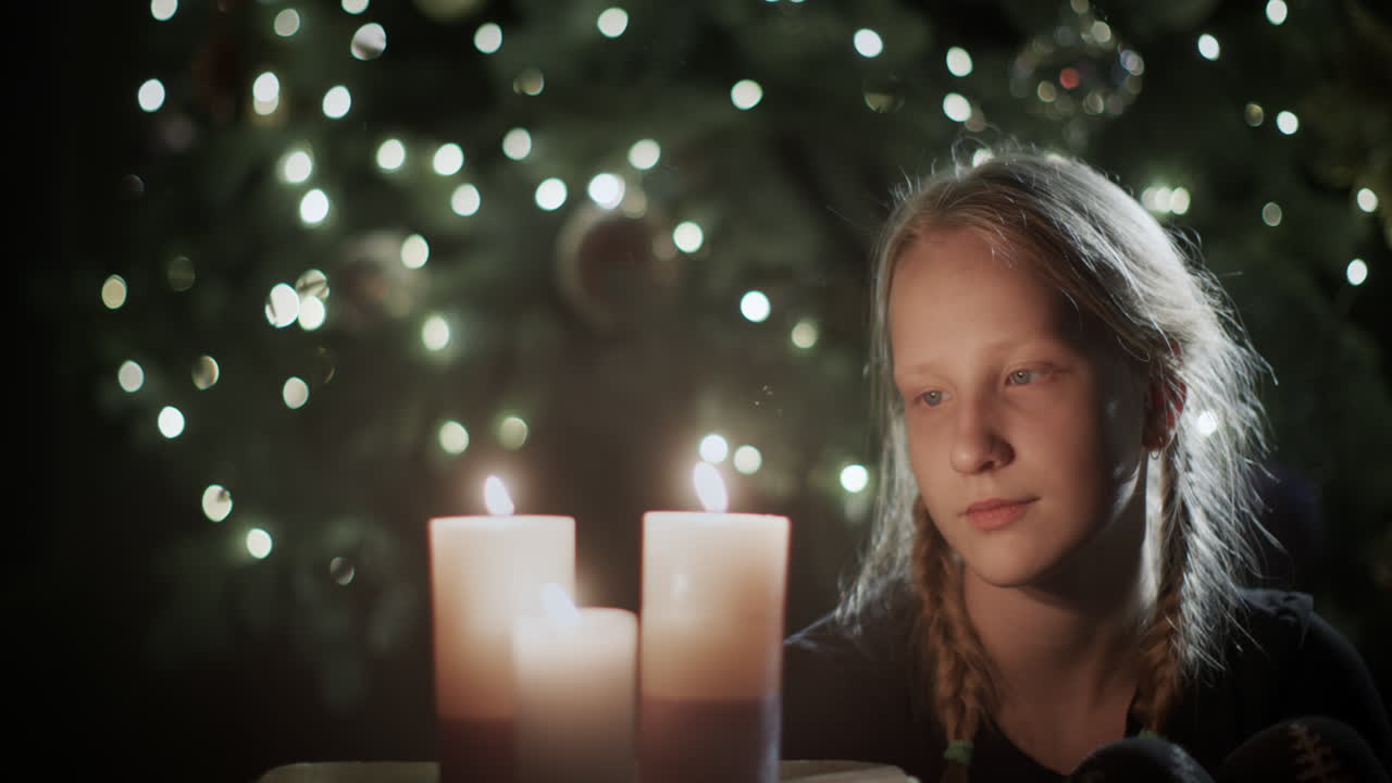 The girl looks at the burning candles. In the background is a beautiful Christmas tree. Children's dreams on Christmas Eve
