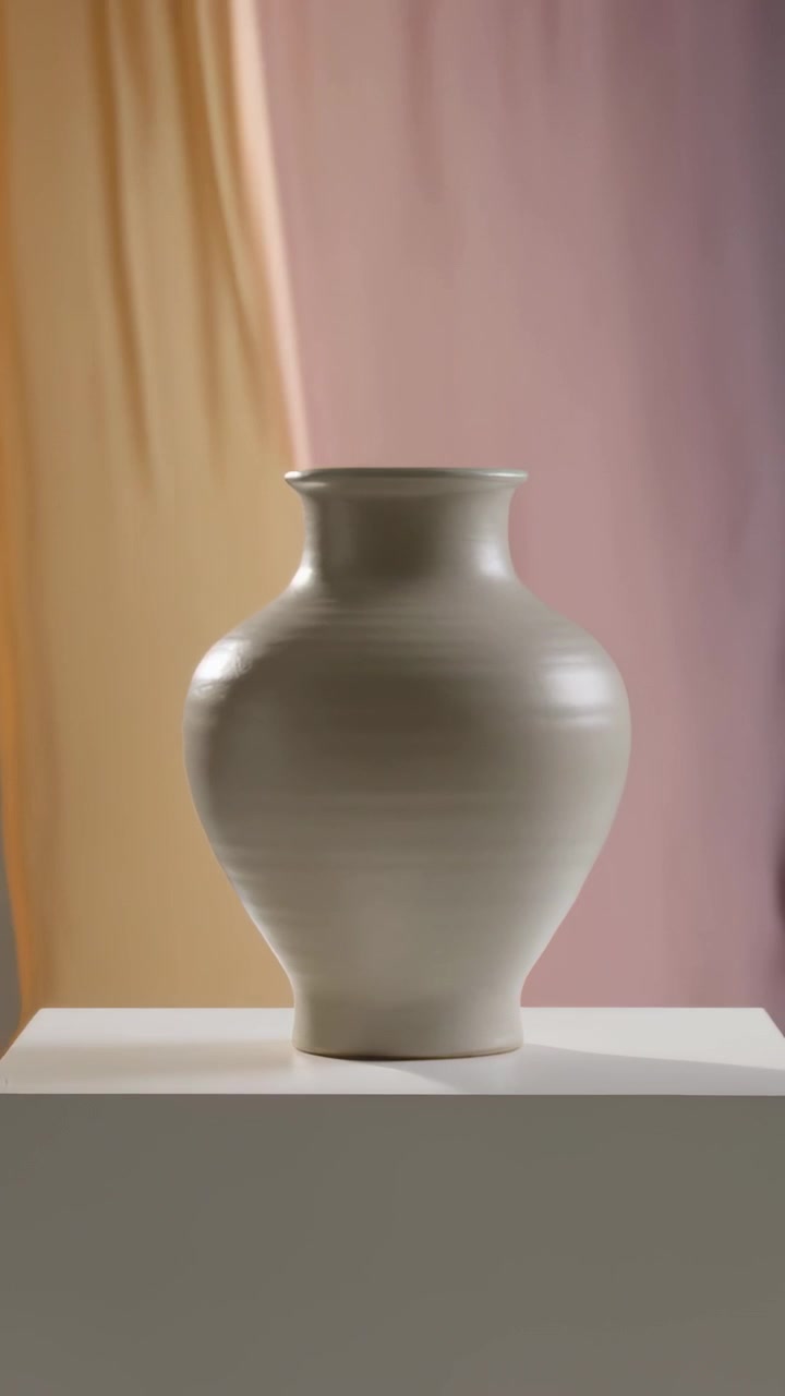 Vertikal video: Kamera trækker sig tilbage og viser en keramisk vase på en hvid sokkel med stofdrapering i et studie.