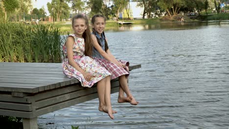 two happy little girls are sitting in the summer on a wooden bridge by the river in the city park next to each other with their bare feet over the water. they look at the camera and laugh.