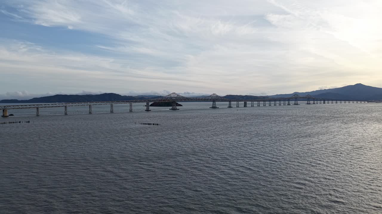 A wide aerial view shows the full reach of the Richmond–San Rafael Bridge amid clouds, cars, and waves in motion.