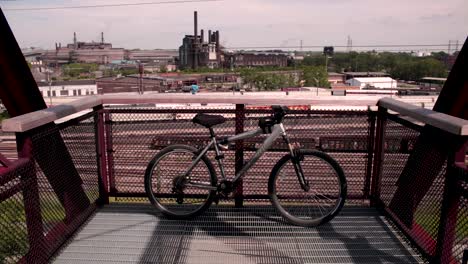 Una-Bicicleta-Permanece-Inactiva-A-Lo-Largo-Del-Borde-De-Un-Puente-Con-La-Industria-Al-Fondo