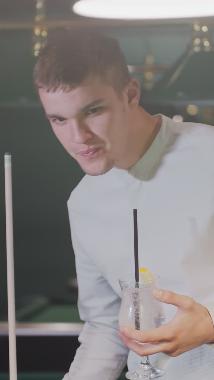 jugador de billar con camisa blanca y pantalones grises sostiene el palo de la pilla y beba una bebida de limón mientras camina en una sala de billar débilmente iluminada. las mesas de billar verdes y las lámparas aéreas crean un entorno atmosférico