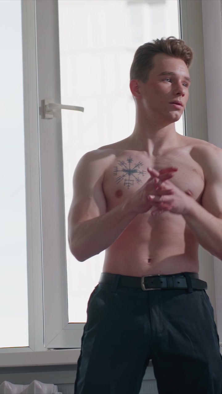 Giovane uomo a torso nudo con tatuaggio geometrico sul petto esegue movimenti con le braccia. L'uomo alza e abbassa le braccia e batte le mani. La scena è ambientata all'interno vicino alla luce del giorno che filtra da una finestra.