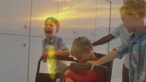 animation of sunset landscape over happy diverse schoolboys pushing friend in wheelchair