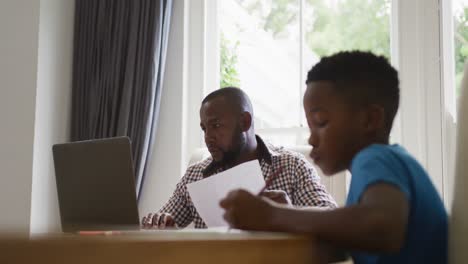 hombre afroamericano trabajando en casa usando una computadora portátil, sentado en la mesa con su hijo haciendo tareas escolares