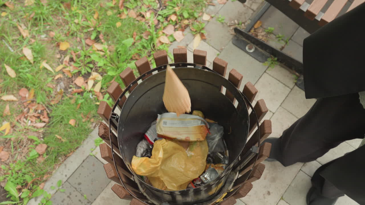 Overhead park bin filled with litter. Person standing beside metal container on paved pathway, black trousers and shoes visible. Discarded wrappers, plastic bag, paper scraps inside bin. Fallen leaves