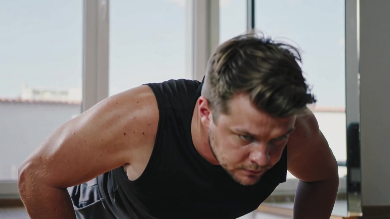 Starting low-angle camera, man doing pushups on hardwood floor training in black tanktop and shorts