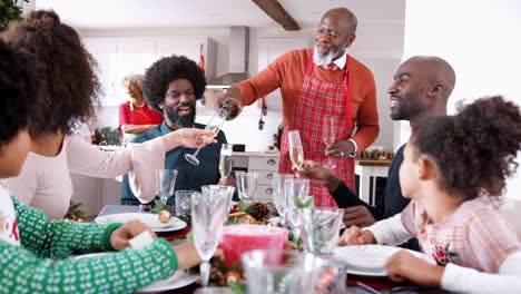 Senior-black-man-wearing-an-apron-pours-champagne-for-his-family-to-celebrate-at-the-dining-table-while-Christmas-dinner-is-prepared,-selective-focus