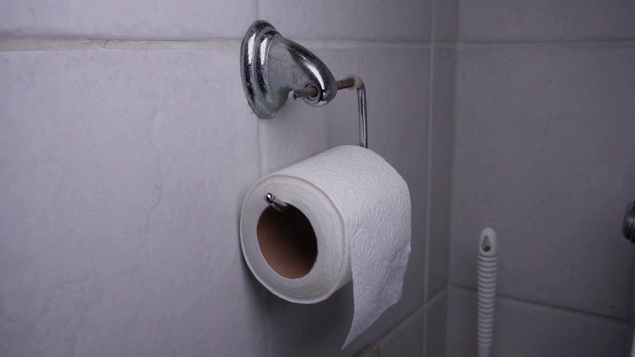 A hand places a roll of toilet paper onto a metal toilet paper holder mounted on a bathroom wall.