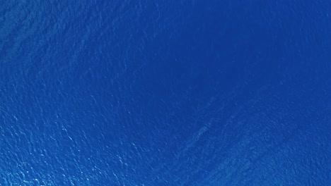 Drone-top-down-bird's-eye-view-moves-across-blue-ocean-water-view-from-surface-as-ripples-and-waves-blow-across-top,-ocean-texture