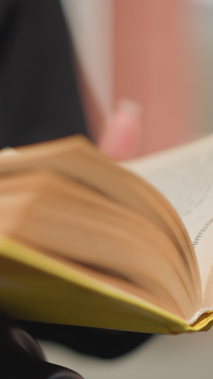 Close-up of book pages being turned by a woman with well-manicured nails and adorned with a ring, subtle blurred background enhancing focus on the text