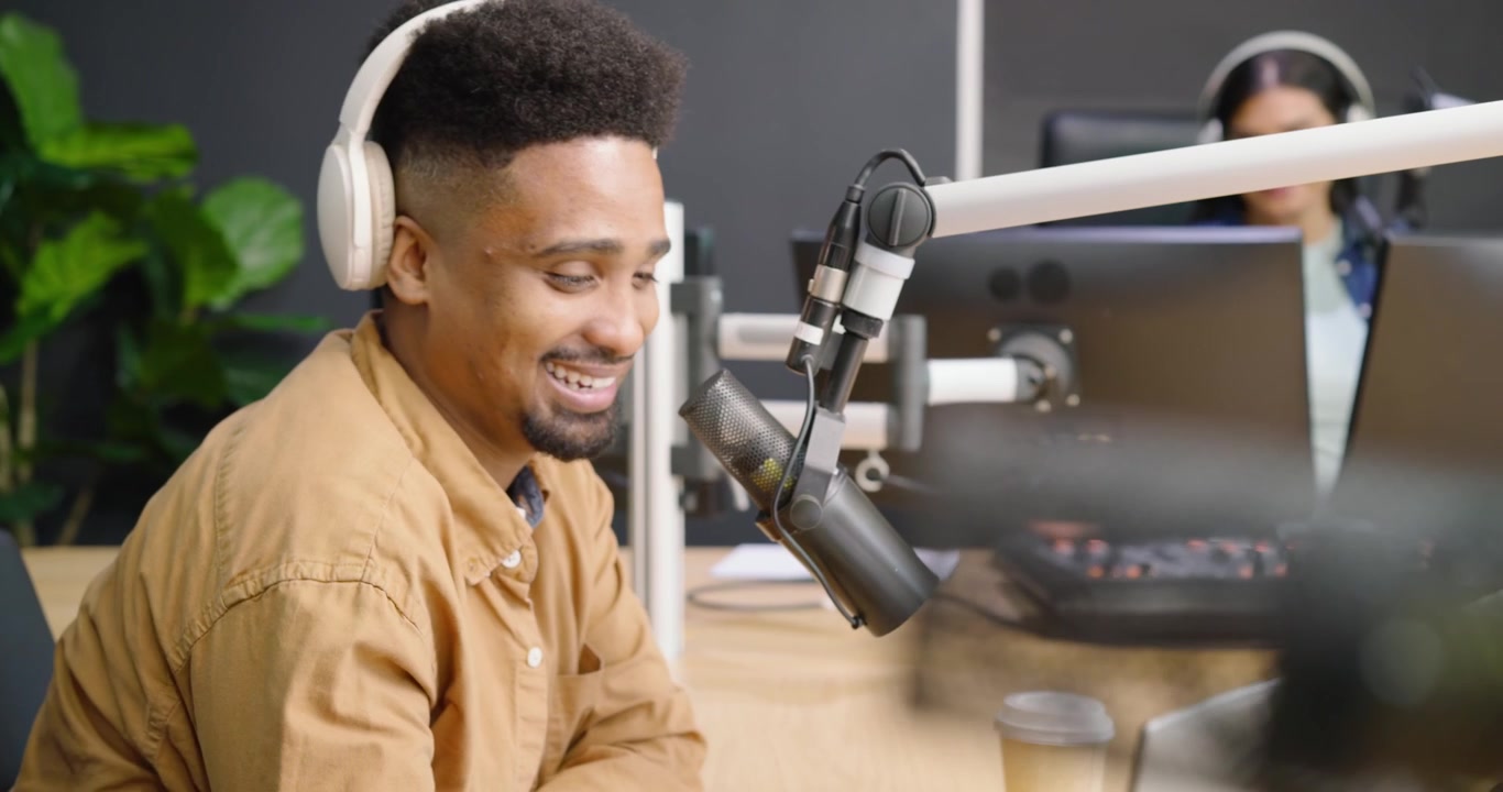 Male host leaning into mic, responding to female host gesturing, using laptop and cup in studio