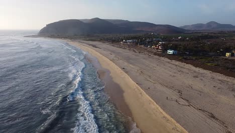 Menschen-Gehen-Bei-Sonnenuntergang-Am-Strand-Spazieren,-Während-Die-Wellen-Gegen-Die-Sandküste-Mit-Bergen-In-Der-Nähe-Von-El-Pescadero-Krachen