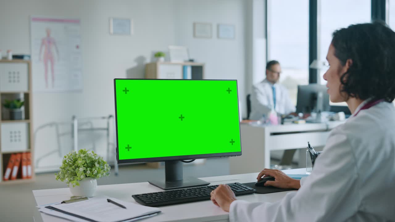 여성 의사는 건강 클리닉에서 녹색 화면 모의 디스플레이를 가진 컴퓨터에서 일하고 있습니다. 색 실험실 코트를 입은 보좌관은 병원 사무실의 책상 뒤에서 의학 역사를 읽고 있습니다.