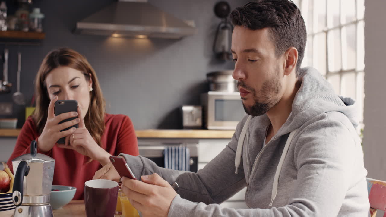 Couple using smart phone devices eating breakfast at home drinking coffee