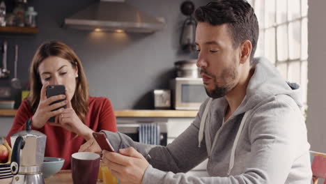 Couple-using-smart-phone-devices-eating-breakfast-at-home-drinking-coffee