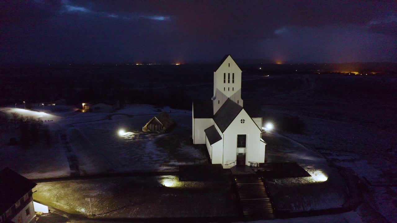 Drone footage capturing a hauntingly beautiful Icelandic church at night, tucked in a quiet snow-dusted village beneath dark northern skies.