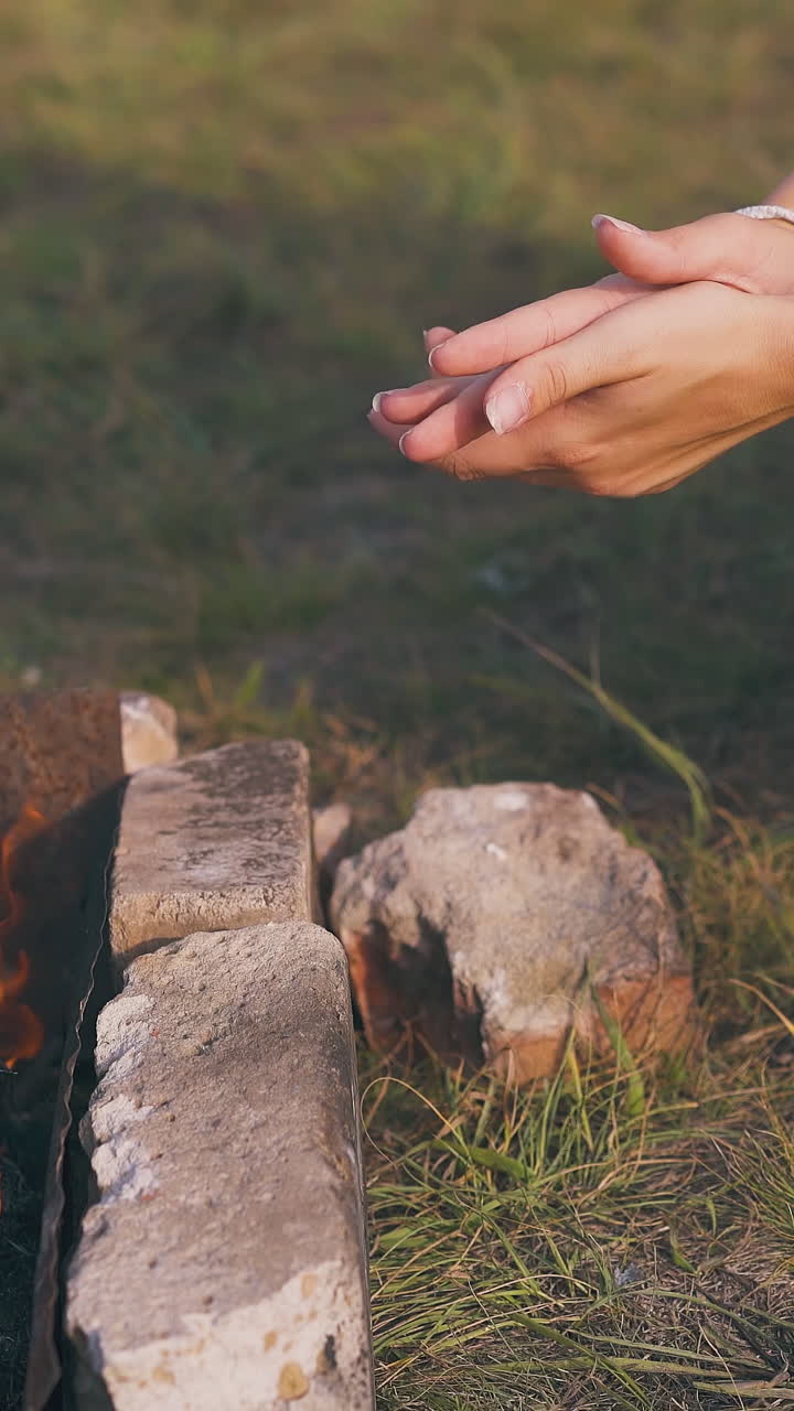 girl hiker in silver jacket heats hands at burning bonfire on meadow at camp in autumn morning closeup