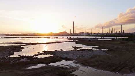 aerial dolly above asphalt lake and white piles of refuse at sunset with oil refinery stacks in distance