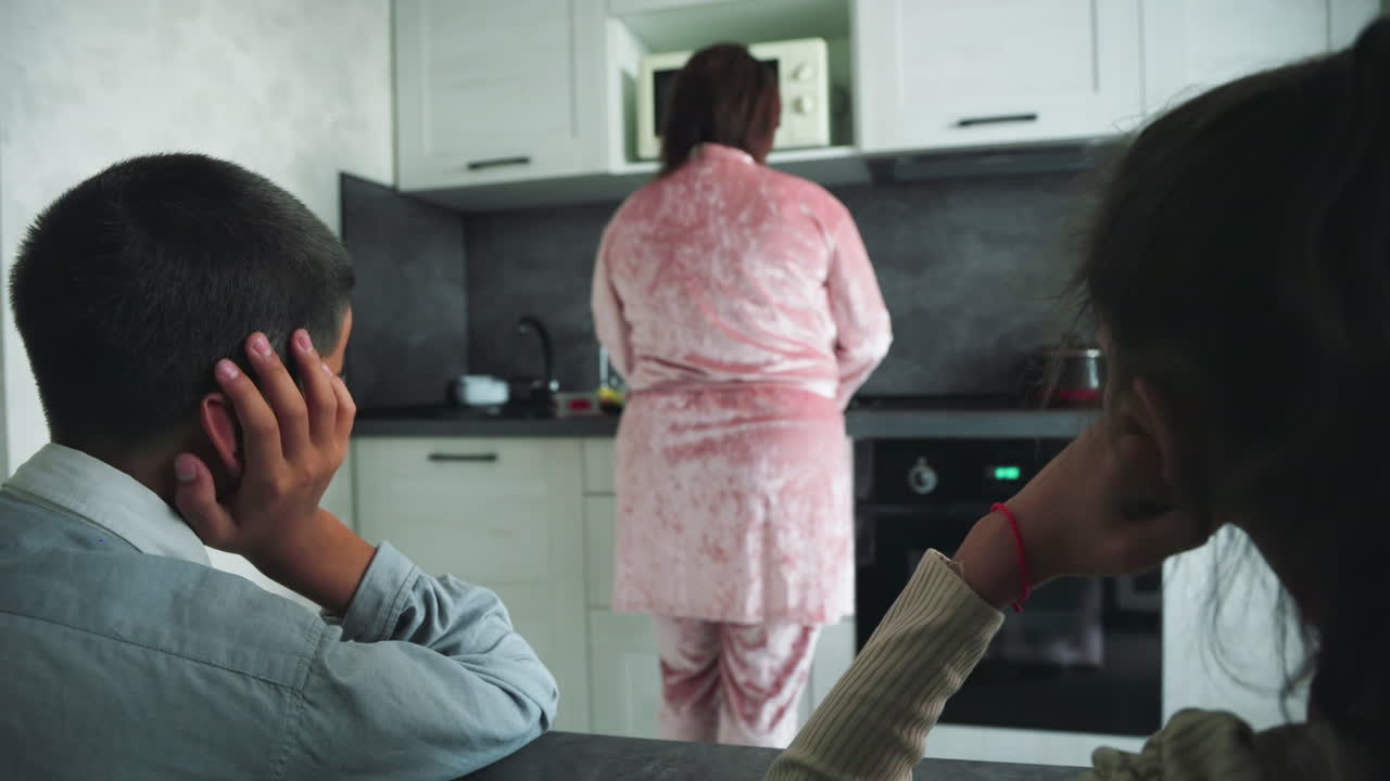 Boy resting head on hand stares toward woman in pink robe preparing food in modern kitchen, microwave above stove, early morning atmosphere, partial view of girl on right, calm household routine