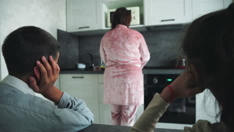 boy resting head on hand stares toward woman in pink robe preparing food in modern kitchen, microwave above stove, early morning atmosphere, partial view of girl on right, calm household routine