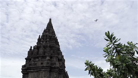 indonesia,prambanan temple on a cloudy day,a flight passes by from the top of the temple.camera tilts down slowly to the ground level, people walking around the temple complex, some posing.