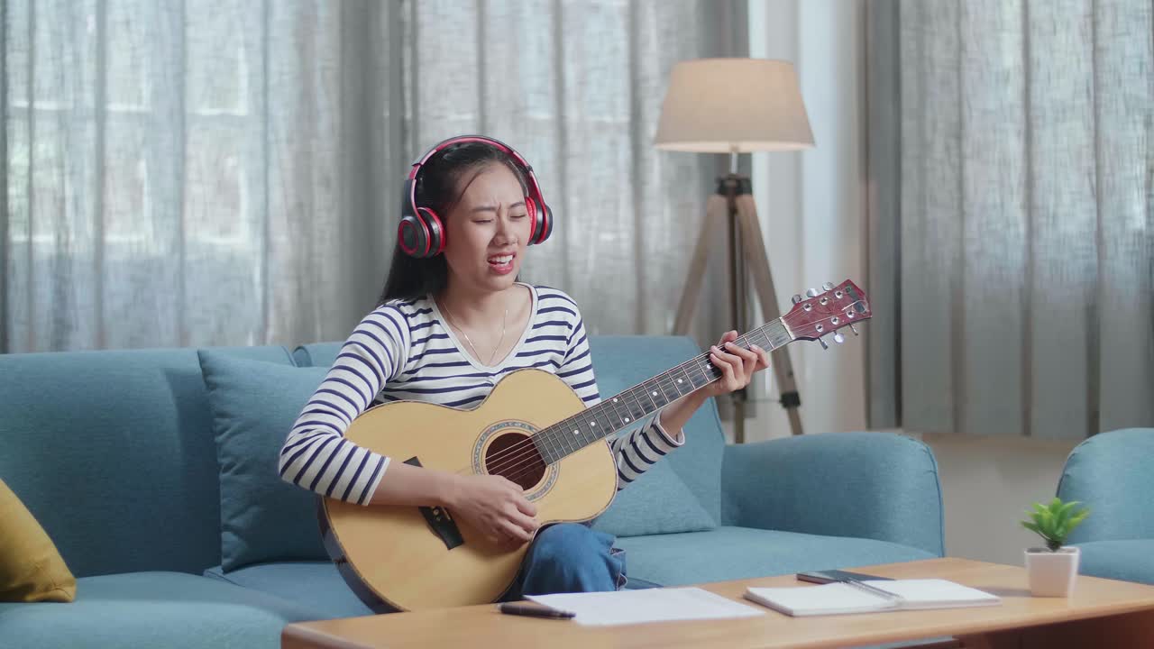 아시아 여성 작곡가가 책상 위에 종이와 노트북을 들고 헤드폰을 착용하고 집에서 노래하고 기타를 연주하고 있습니다.