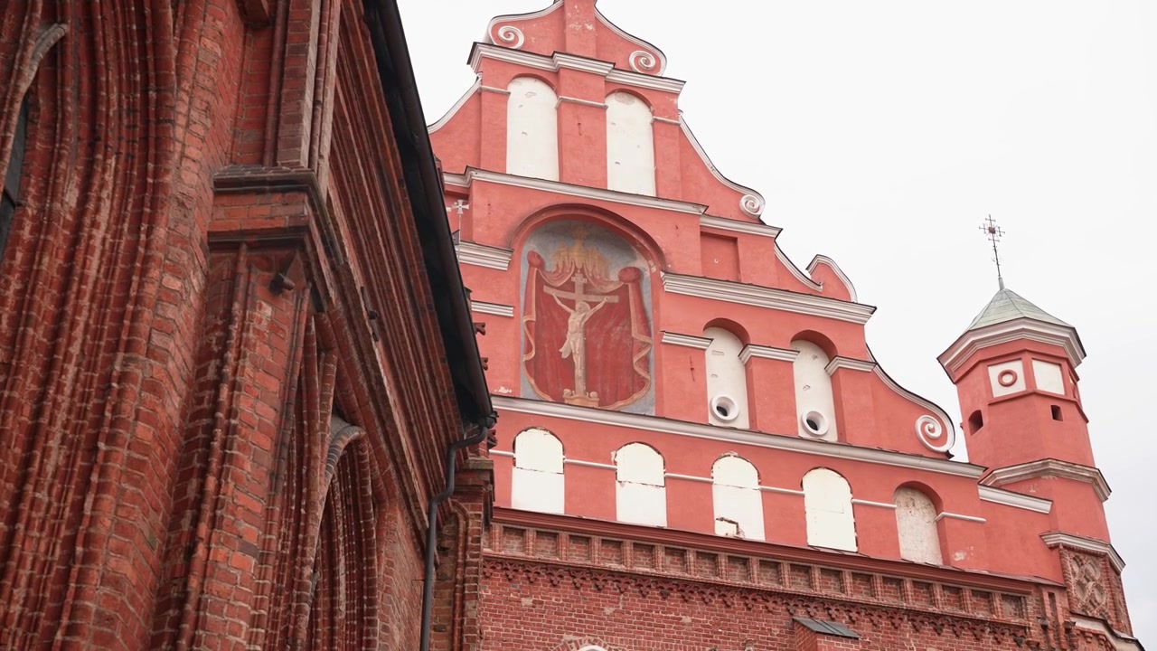 Inquadratura grandangolare della Chiesa di San Francesco e San Bernardino a Vilnius, Lituania. Mostra una facciata ornata in mattoni rossi, luce soffusa e nuvolosa, architettura gotica e rinascimentale, nessuna persona visibile.