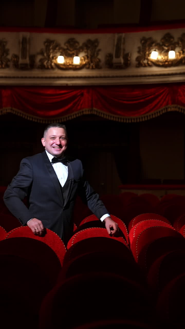 Man stands alone among velevet chair in classic theater. Male in classic suit invites public come in with gesture. Time before performance. Vertical video
