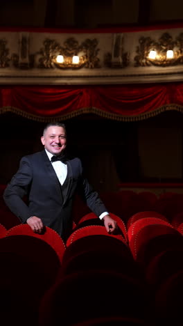 man stands alone among velevet chair in classic theater. male in classic suit invites public come in with gesture. time before performance. vertical video