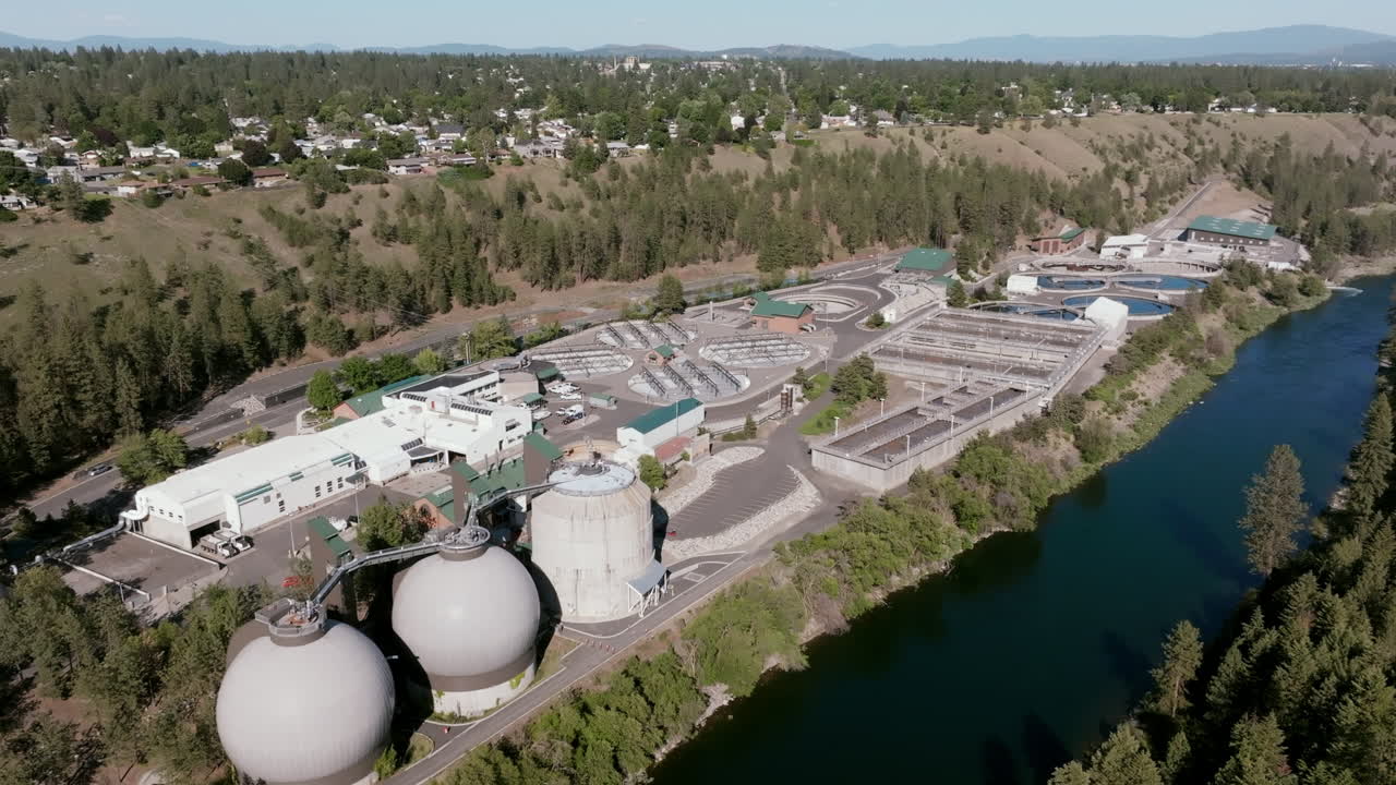 A wide aerial shot of the Spokane water treatment plant shows its complex layout along a forested hillside and river, with nearby residential areas and mountains in the distance