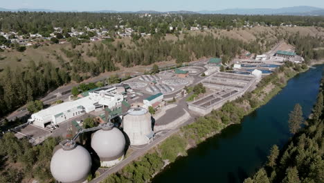 a wide aerial shot of the spokane water treatment plant shows its complex layout along a forested hillside and river, with nearby residential areas and mountains in the distance