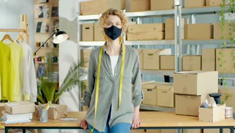 young caucasian woman in mask looking at camera in good mood sitting on table in own shop with clothes