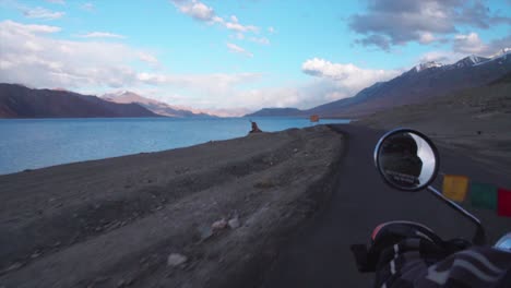 driving slowly with a motorbike along a lake in the himalaya moutains at sunset with pink clouds and blue skies.
