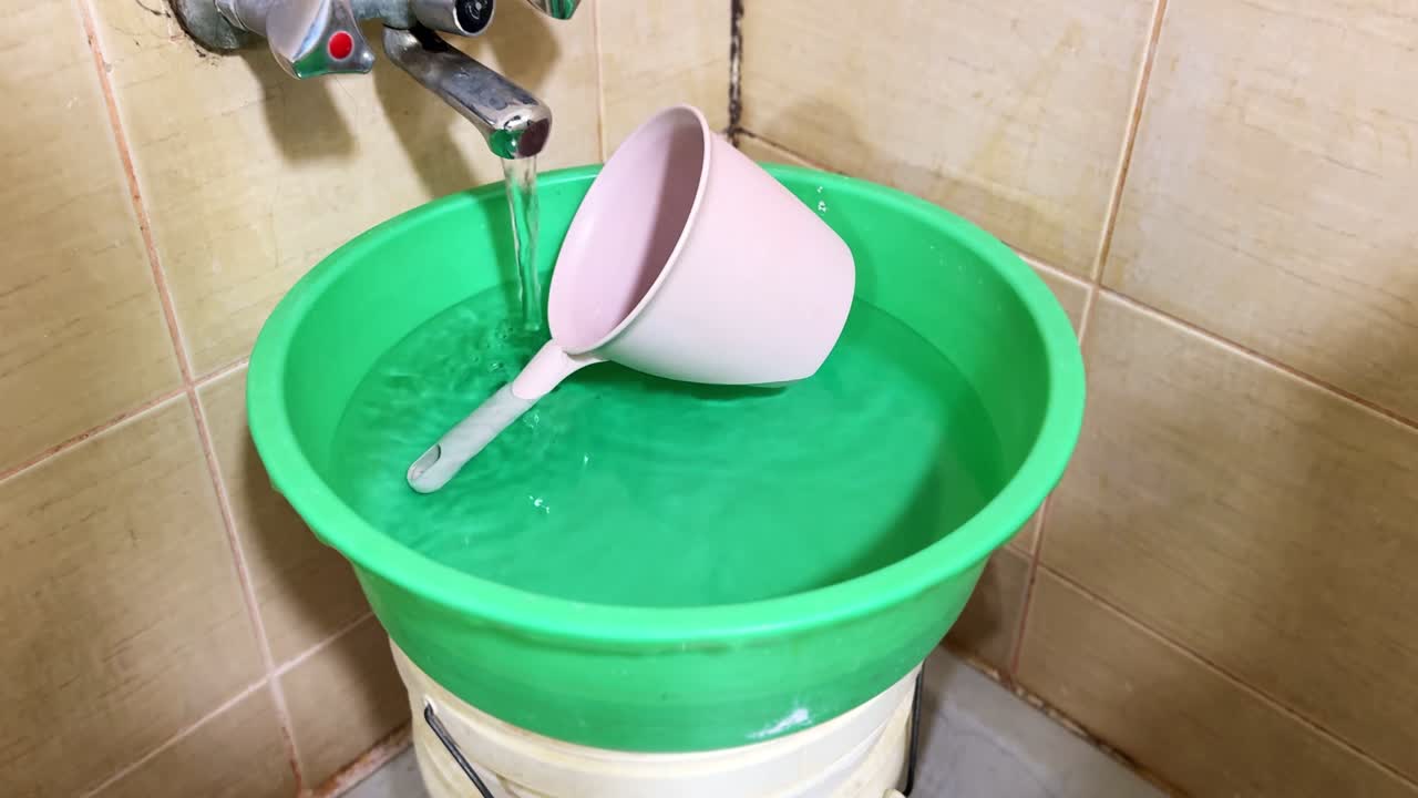 Close-up of running faucet pouring into a green plastic basin with pink dipper in tiled bathroom corner, indoor daylight lighting, household cleaning and hygiene concept