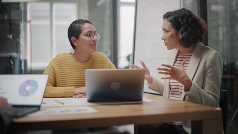 dos mujeres de diversas etnias tienen una discusión en una sala de reuniones detrás de paredes de vidrio en una agencia. el director creativo y el gerente de proyecto comparan los resultados comerciales en diseños de portátiles y aplicaciones en una oficina.