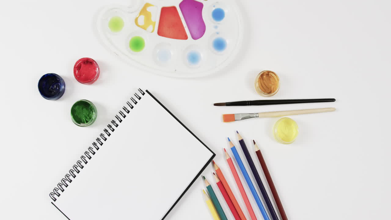 vista aérea del cuaderno de notas, pinturas, pinceles y lápices de colores sobre fondo blanco, en cámara lenta