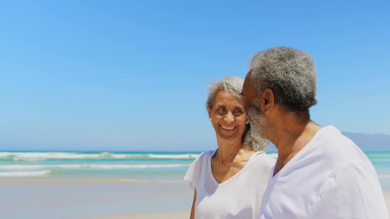 Side view of happy active senior African American couple walking on beach in the sunshine 4k