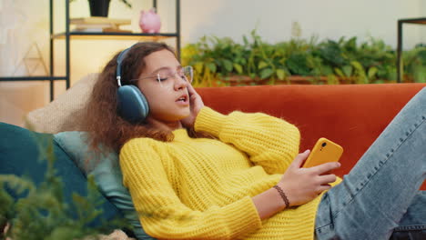 Happy-teen-girl-in-wireless-headphones-relaxing-sitting-on-sofa-at-home-listening-favorite-music