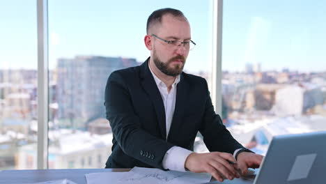 serious business worker on office window backdrop. middle aged businessman in glasses sitting at desk in his workplace. entrepreneur searches solution of business issues.