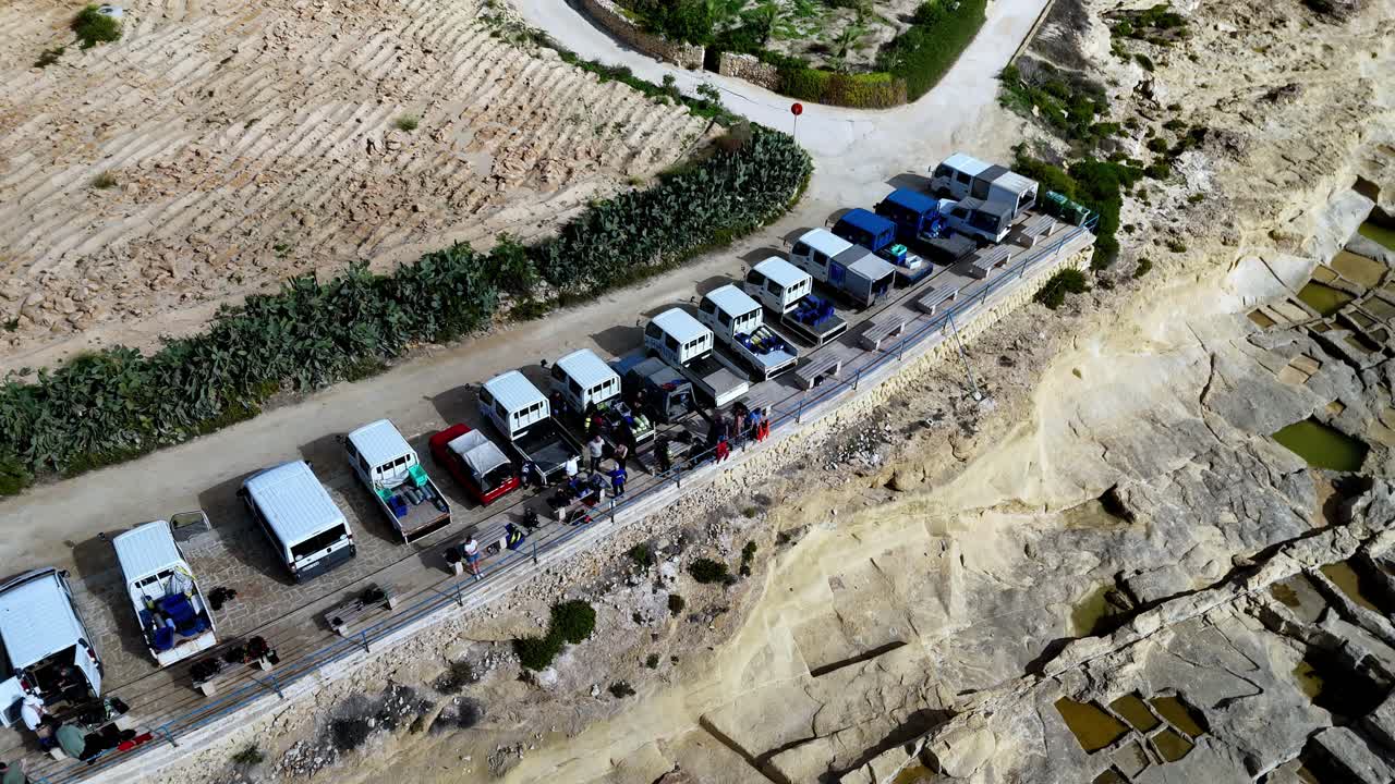 Aerial drone shot of diverse diving expedition trucks parked on Gozo limestone cliffs during a sunny Mediterranean summer day featuring vibrant blue and white vehicles against rugged coastal terrain
