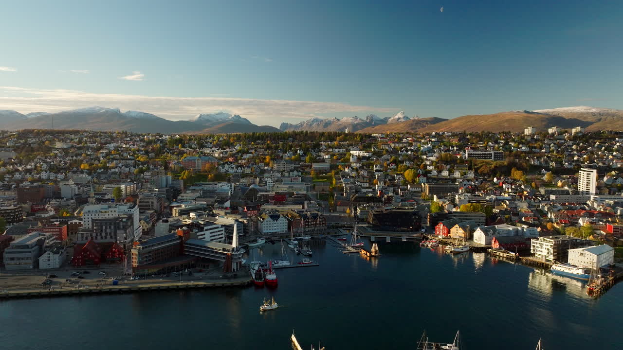 Aerial drone view of Tromso, Norway in late autumn. The city center and harbor are illuminated by low sun, with snow-capped mountains in the background. A scenic Arctic landscape