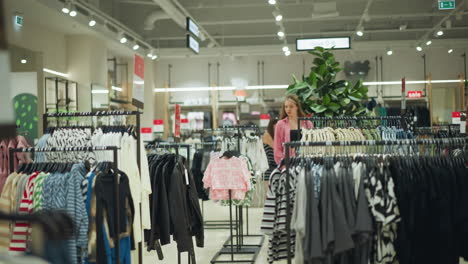 lady wearing black crop top under pink outfit carries shopping bag while walking through a well-lit shopping mall filled with neatly arranged clothes on racks, she explores the fashion section